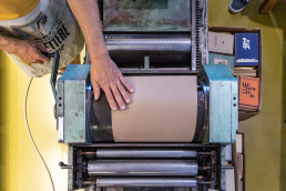 A person operates a vintage printing press with pink paper, surrounded by colorful books in a workshop setting.