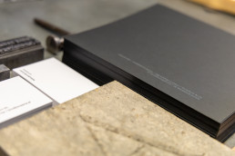 Portfolio mockup showing dark grey branded materials and white business cards on textured beige surface for Enaena project