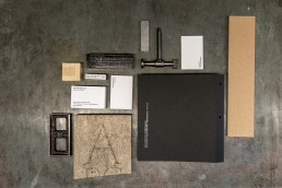 Flat lay mockup showing Enaena branding materials including black folders, kraft paper, white business cards, and vintage pri