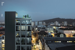 Modern dark residential tower with illuminated windows at dusk overlooking urban cityscape with hills in background.