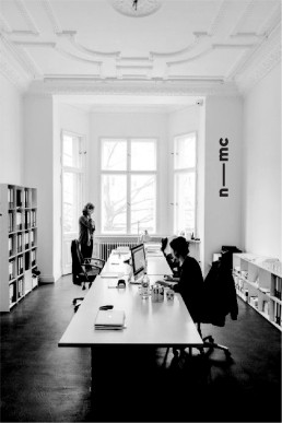 Black and white photo of nmc office interior with ornate ceiling, bookshelves, long white desk, and people working at laptops