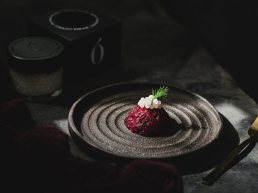 Fine dining presentation on dark textured plate with beetroot dish, white garnish and dill, moody lighting with teapot in bac