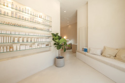 Modern minimalist retail interior with glass shelving displaying white skincare products, cream seating area, and potted plan