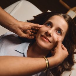 Young woman with long brown hair lying on white bedding, hands framed around her face, wearing light blue top with pearl brac