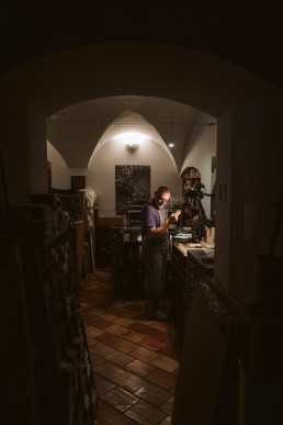 Workshop scene showing an older man in purple shirt working at vintage printing press in atmospheric arched stone interior wi