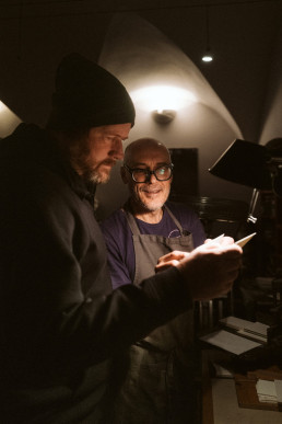 Two chefs in dark aprons collaborating in dimly lit kitchen, one wearing beanie, other with glasses, reviewing notes together