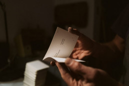 Hands holding an open white document or brochure with text, photographed in warm natural lighting with soft shadows