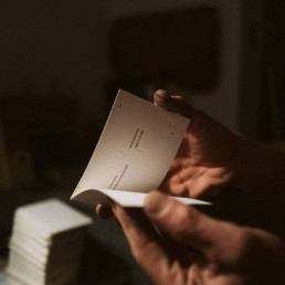 Hands holding an open white document or brochure with text, photographed in warm natural lighting with soft shadows