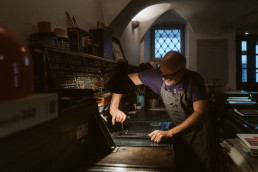 Man operating vintage letterpress printing machine in atmospheric workshop with arched windows and warm lighting