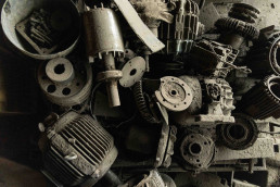 A collection of dusty, weathered mechanical engine parts and components arranged together, showing gears, cylinders, and meta
