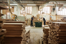 Woodworking workshop with two craftsmen operating machinery among stacked lumber, featuring exposed ductwork and natural ligh