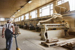 Woodworking workshop with industrial machinery, dust collection system, stacked lumber, and worker in overalls operating equi