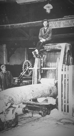 Black and white photograph showing workers operating a vintage sawmill with large mechanical saw and log, industrial machiner