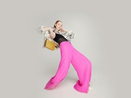 Fashion model in bright pink wide-leg pants, black sheer top, patterned jacket holding yellow quilted bag against neutral bac