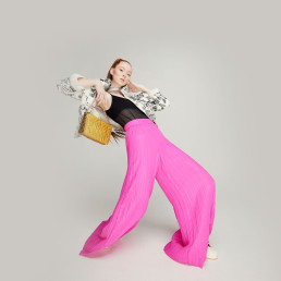 Fashion model in bright pink wide-leg pants, black sheer top, patterned jacket holding yellow quilted bag against neutral bac