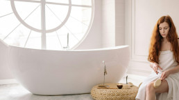 Mockup of woman in white towel sitting beside modern white bathtub in bright bathroom with circular window for Hello universe