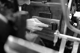 Black and white photograph showing hands operating vintage printing press machinery, with focus on traditional typesetting eq