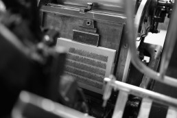 Black and white close-up of vintage printing press machinery with printed text page being processed through metal mechanical