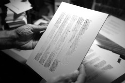 Black and white photograph showing hands holding printed documents with columns of text, with stacks of papers visible in the
