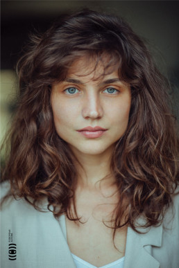 Portrait of a woman with wavy brown hair and blue eyes wearing a light blue top, shot in natural lighting with soft focus bac