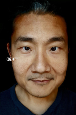 Close-up portrait of an Asian man with dark hair and subtle facial hair wearing a navy blue shirt against black background