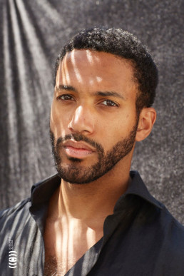 Professional headshot of a man with curly hair and beard in dark shirt against textured grey background with dramatic lightin
