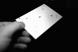 Black and white photograph showing a hand holding a white card or paper with minimal black typography against a dark backgrou