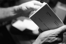 Black and white photo showing hands holding an open book with text on the page, creating an intimate reading moment.